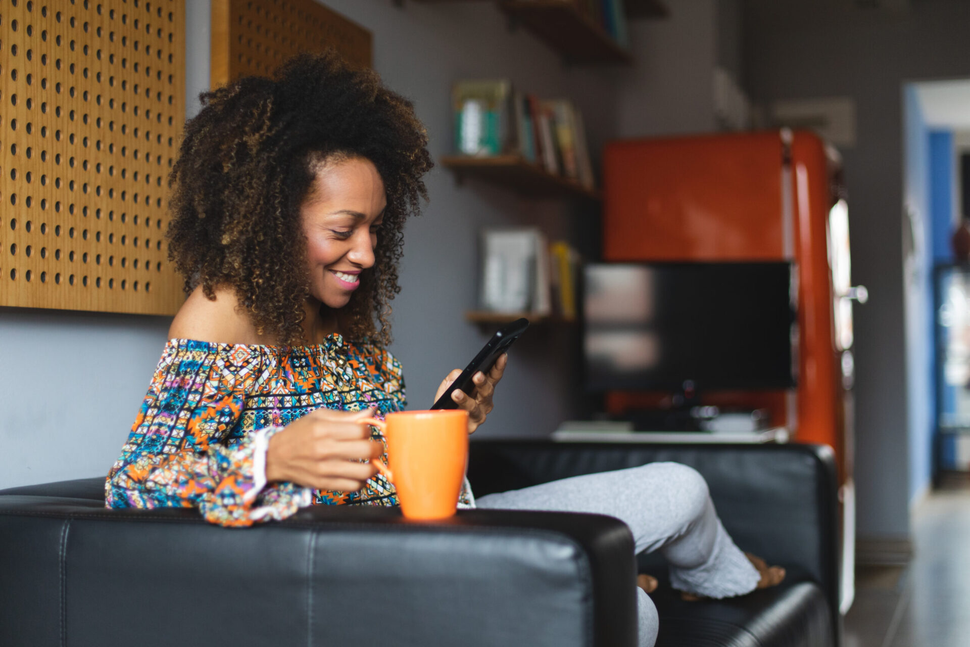 Casual young black woman relaxing at home texting on smartphone and drinking coffeee. Home leisure and tranquility concept. Afro hairstyle model.