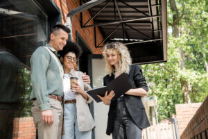 smiling real estate agent showing documents to interracial couple with paper cup near cottage