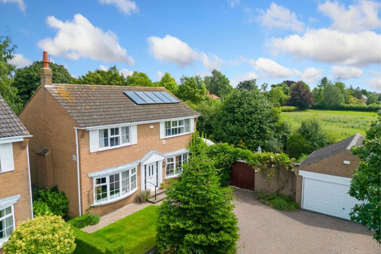Modern two-storey detached house with solar panels, manicured front garden, large driveway, and open countryside in the background under a blue sky.