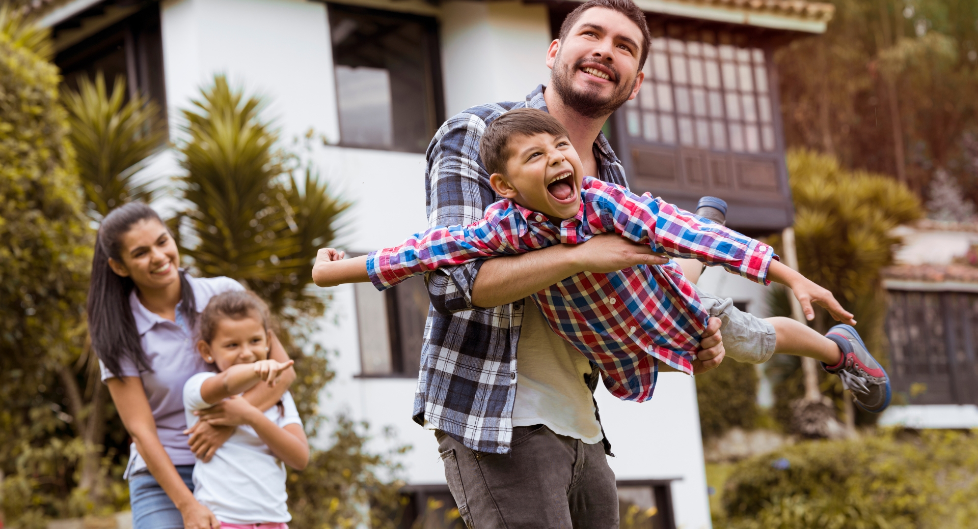 A smiling family with two children playing outdoors in front of their home, representing the importance of researching a neighbourhood before buying a property.