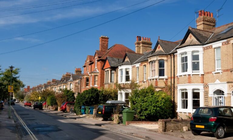 A row of traditional UK terraced houses on a residential street with parked cars and blue sky.