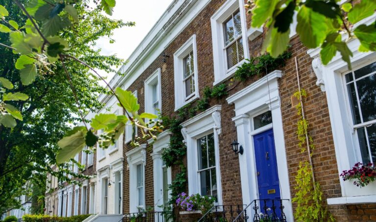 Row of UK terraced rental homes with brick façades, white sash windows, leafy trees, and a blue front door, representing residential rental property pricing in 2026.