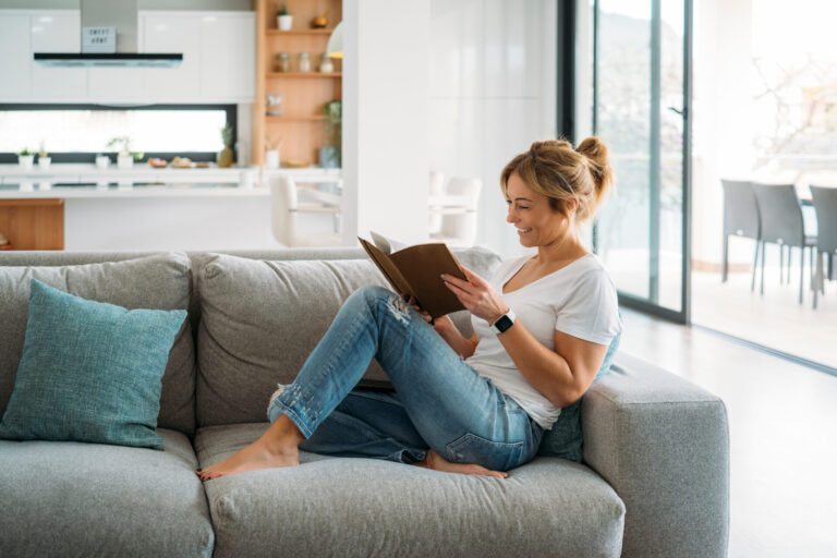Woman relaxing on a sofa reading a book in a bright modern home, representing the peaceful lifestyle and residential appeal of Boley Park WS14.