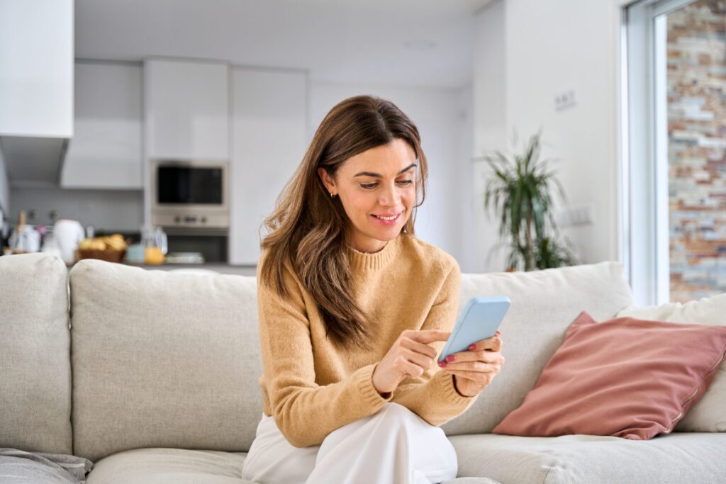 Smiling woman using her phone on a cream sofa in a bright open-plan home, representing living in Fulford YO10, York