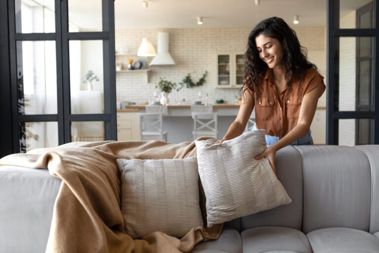 Woman arranging cushions on a grey sofa in a bright open-plan living room and kitchen, representing homes in Oatlands HG2, Harrogate