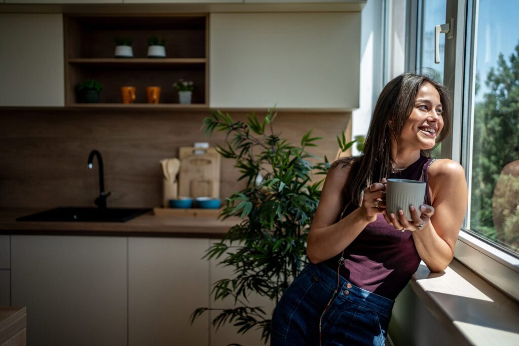 Smiling woman holding a mug of coffee by a sunny kitchen window, representing village living in Powfoot DG12