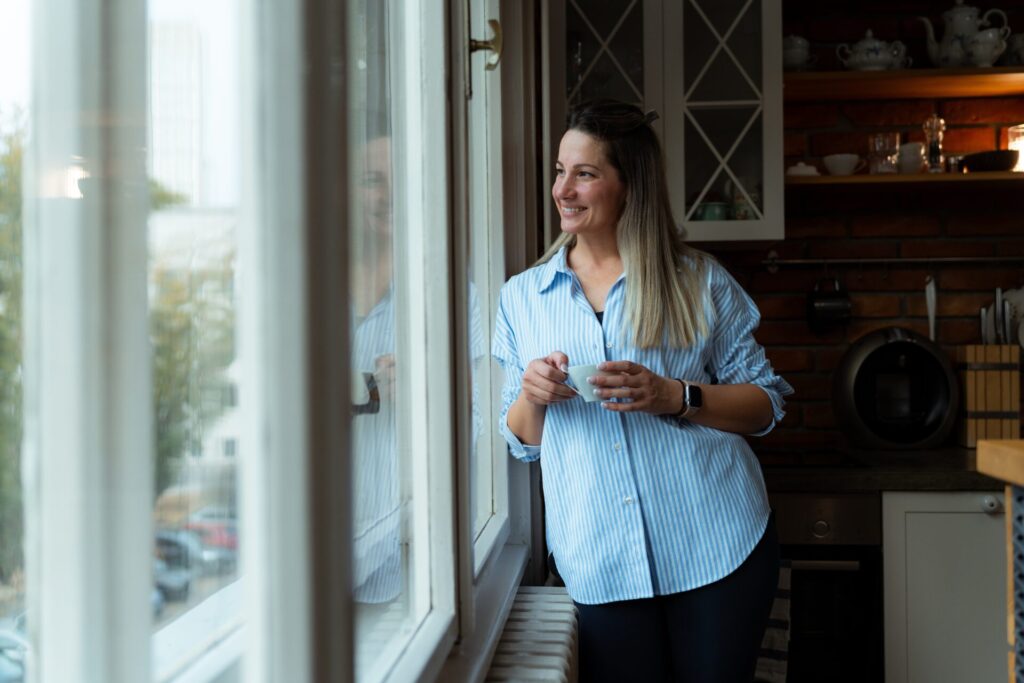 Woman in a striped shirt holding a cup of coffee and looking out of a kitchen window, representing life in Locharbriggs DG1