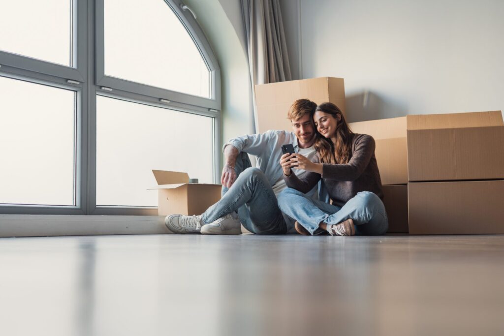 Young couple sitting among moving boxes looking at a phone in their new rental home, representing renting in Menston LS29