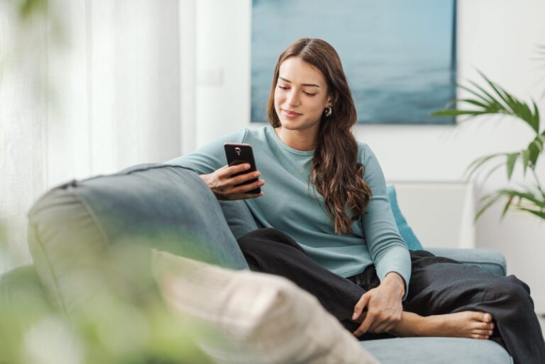 Young woman relaxing on a sofa and using her phone in a bright rental flat, representing renting in Miles Platting M40