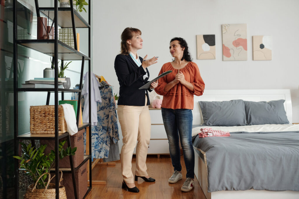Female letting agent holding a clipboard while discussing a rental property's condition with a woman inside a furnished bedroom.
