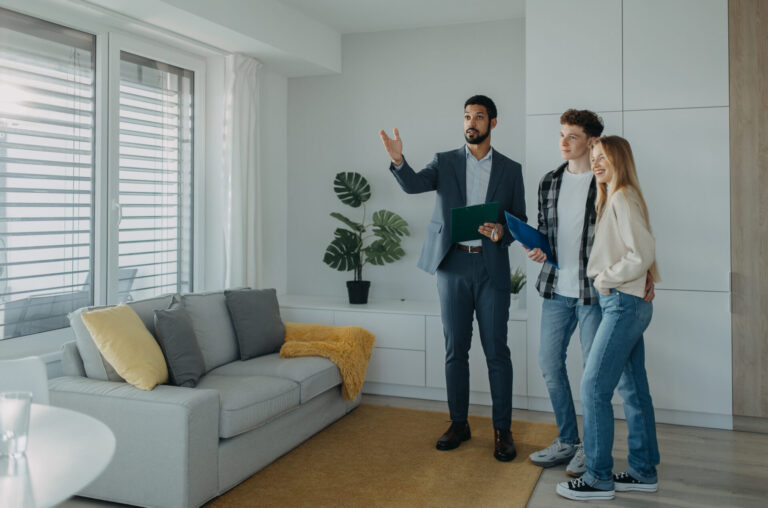 Estate agent showing a bright, modern living room to a young couple during a residential property viewing.