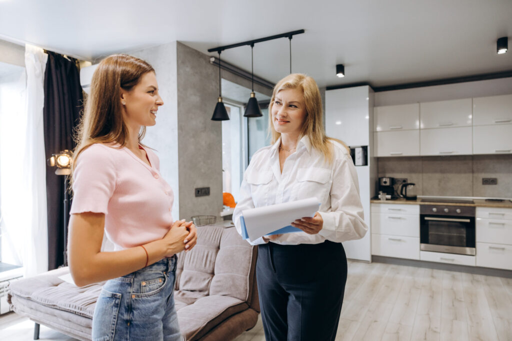 Female estate agent holding property documents while discussing a home with a woman in a bright, modern open-plan living room and kitchen.